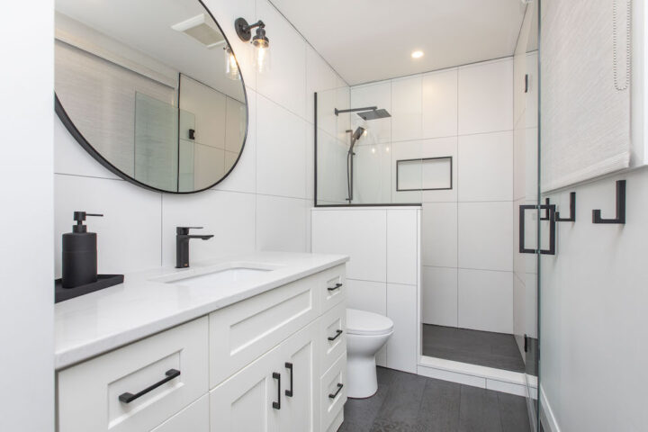 Bathroom with a white shaker vanity, matte black faucet, round black mirror and a glass walk-in shower