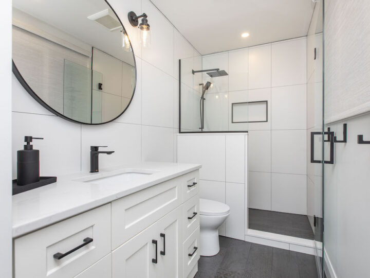 Bathroom with a white shaker vanity, matte black faucet, round black mirror and a glass walk-in shower