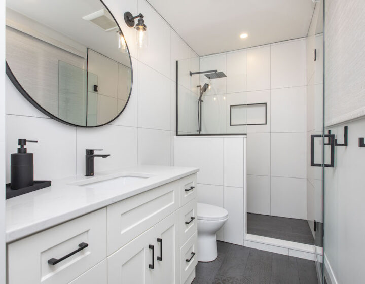 Bathroom with a white shaker vanity, matte black faucet, round black mirror and a glass walk-in shower