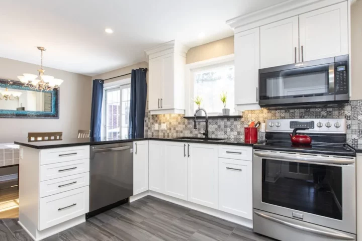 White L-shaped kitchen with black countertops, a patterned tile backsplash and stainless appliances