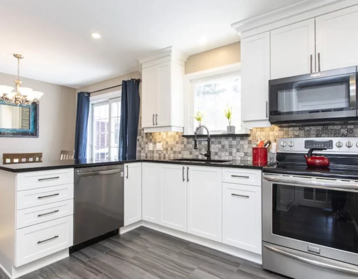 White L-shaped kitchen with black countertops, a patterned tile backsplash and stainless appliances